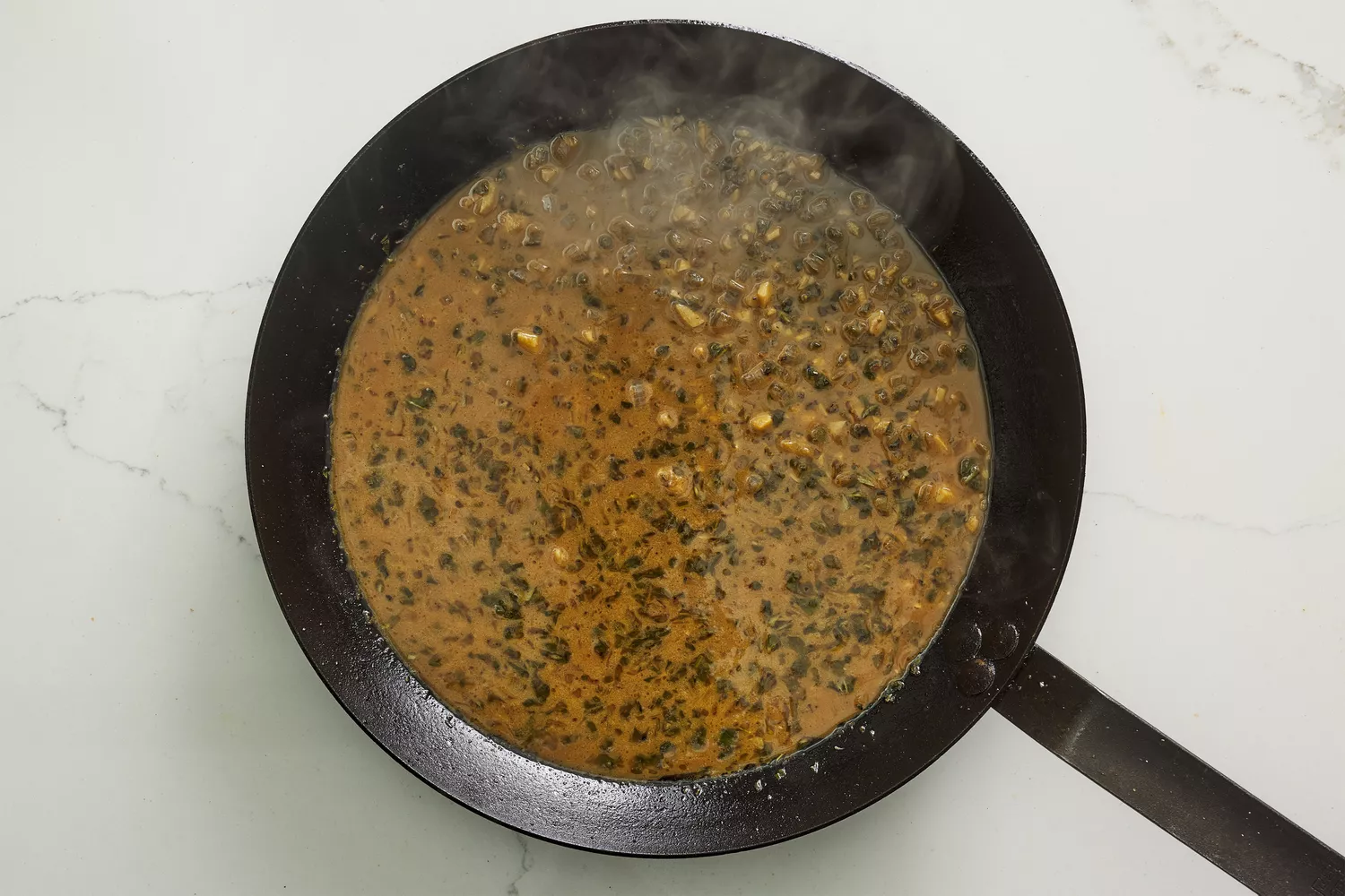A skillet with garlic, herbs, and shallots cooking in a white wine-chicken stock sauce