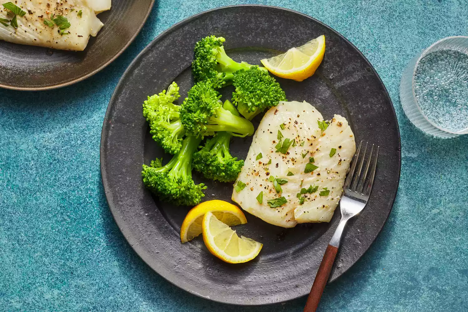 Une assiette de morue au citron et à l'ail cuite au four, servie avec du brocoli vapeur, des quartiers de citron, et un verre de seltzer